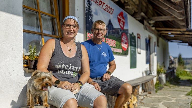 The innkeepers of the Marienseer Schwaig, © Wiener Alpen, Christian Kremsl Two people sitting in front of a building with a dog, in the background a poster with the date Sept. 09, 2023.