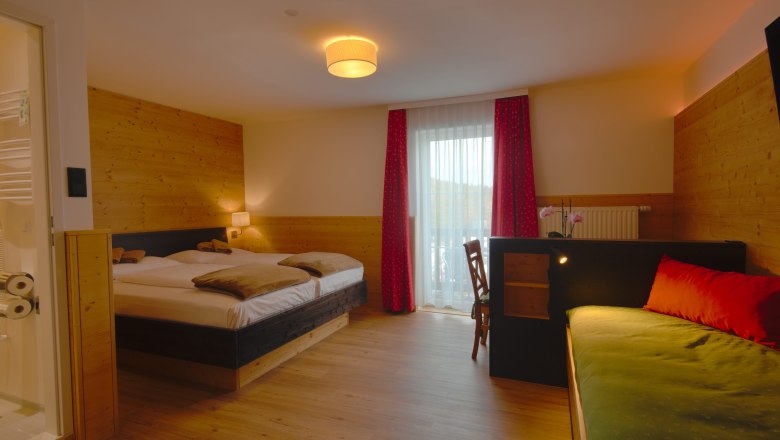 Double room, © Alpengasthof Enzian A cozy double room with wooden walls, a double bed, a sofa and a desk. A window with red curtains lets in natural light.