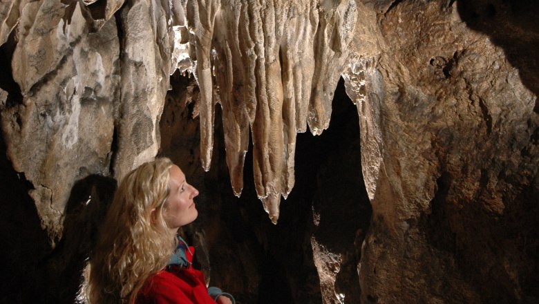 Hermannshöhle, © Hermannshöhle Eine Frau in roter Jacke betrachtet Stalaktiten in einer Höhle.