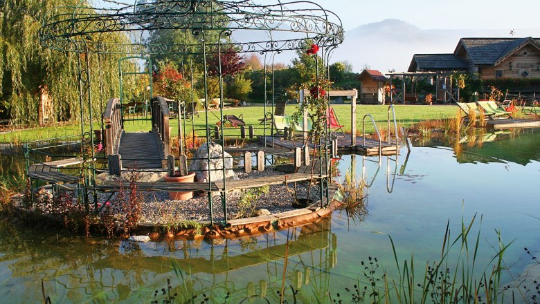 Sinnesfreuden im Paradiesgart'l, © Hotel Molzbachhof, Foto: Familie Pichler Ein idyllischer Garten mit Pavillon, Brücke und Teich, umgeben von grüner Landschaft und Hütten im Hintergrund.