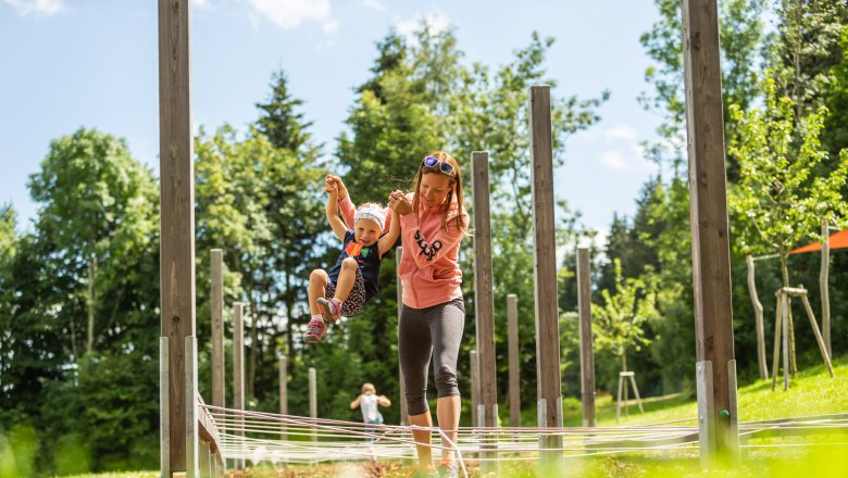 St. Corona motor skills park, © Wexl Arena St. Corona am Wechsel A woman holds a child by the hands in an outdoor play area with wooden posts and ropes.