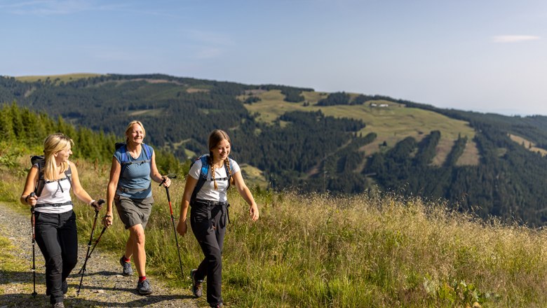 Tip: 3 huts Schwaigen tour, © Wiener Alpen, Christian Kremsl Three women hike along a mountain path with hiking poles and rucksacks, surrounded by green hills and forests.