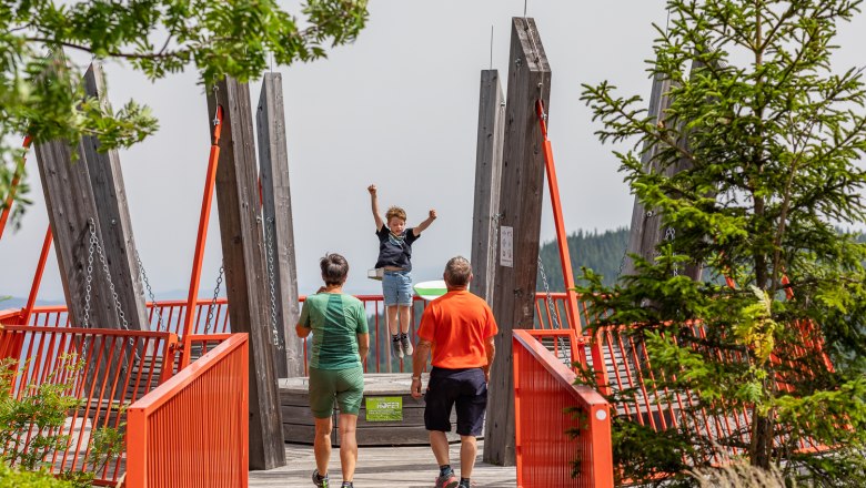 Alpine swing viewing platform, © Wiener Alpen/Kremsl A child swings on a viewing platform with red railings while two adults look on.