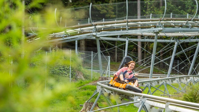 Summer toboggan run, © Erlebnisarena St. Corona A woman and a child ride through a green landscape on a summer toboggan run, laughing.