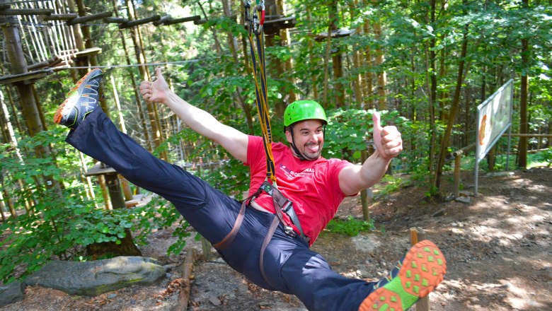 Flyingfox, © Hamari Kletterpark Person mit Helm und Sicherheitsgurt auf einer Seilrutsche im Wald.