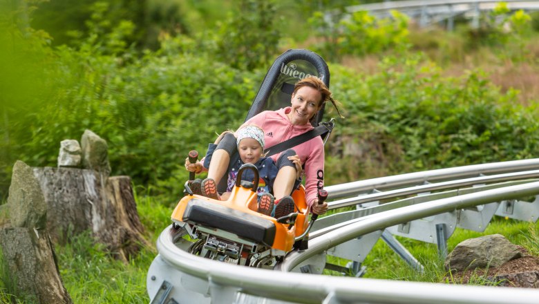Summer toboggan run, © Erlebnisarena St. Corona A woman and a child ride on a summer toboggan run through a green landscape.