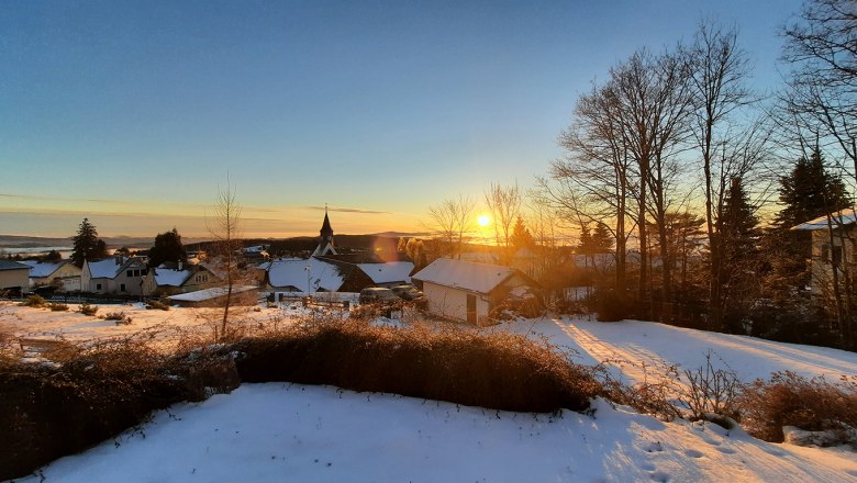 Sonnenaufgangsblick im Winter, © Christoph Gierlinger Winterlandschaft mit Sonnenaufgang über einem verschneiten Dorf und kahlen Bäumen.