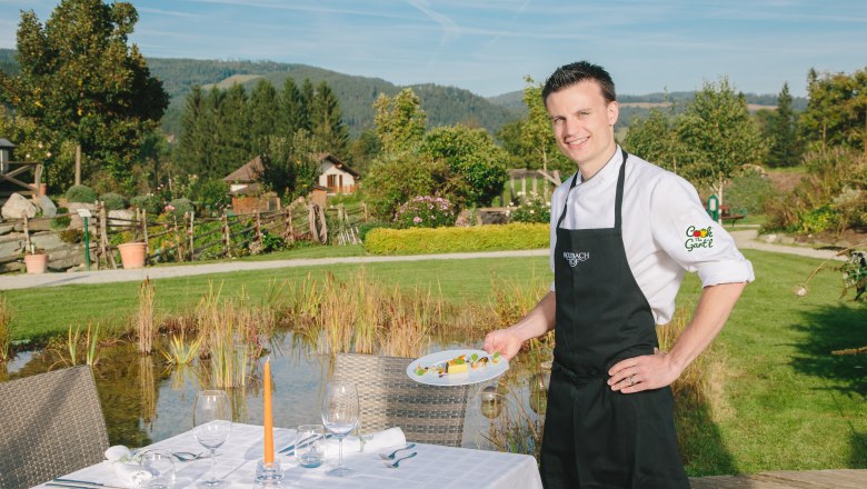 peterpichler_c_molzbachhof, © Hotel Molzbachhof Ein Koch in schwarzer Schürze steht draußen neben einem gedeckten Tisch und hält einen Teller mit Essen. Im Hintergrund ist eine grüne Landschaft zu sehen.