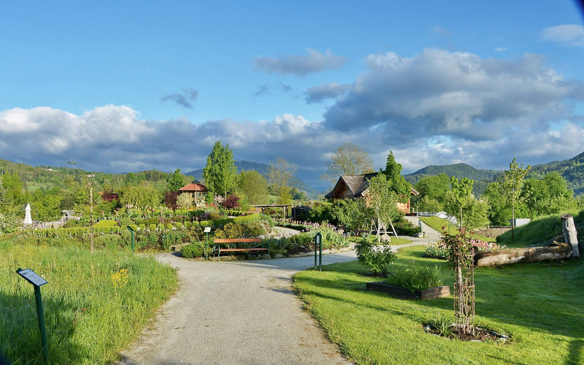 Ein gepflegter Garten mit Wegen, Bäumen und Blumen, umgeben von grünen Hügeln und einem blauen Himmel mit Wolken.