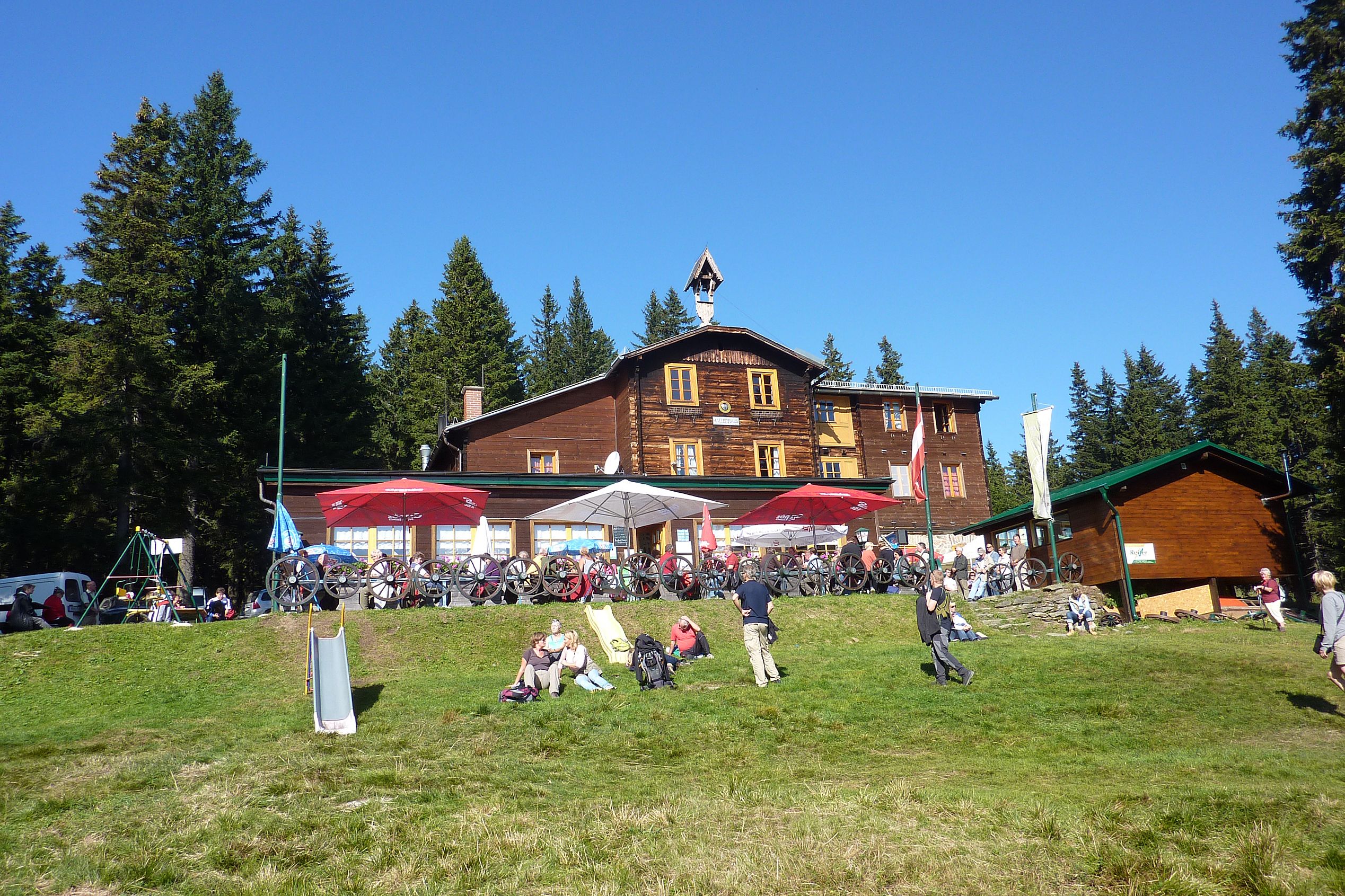 Holzhaus in den Bergen mit Menschen und Fahrrädern davor, umgeben von Bäumen.