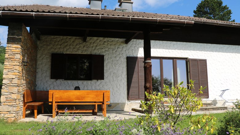 A house with a wooden seating area on the terrace, surrounded by a garden with flowers.