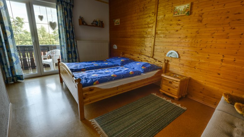 Gemütliches Schlafzimmer mit Holzwänden, Doppelbett, blauem Bettzeug und Balkon mit Blick ins Grüne.