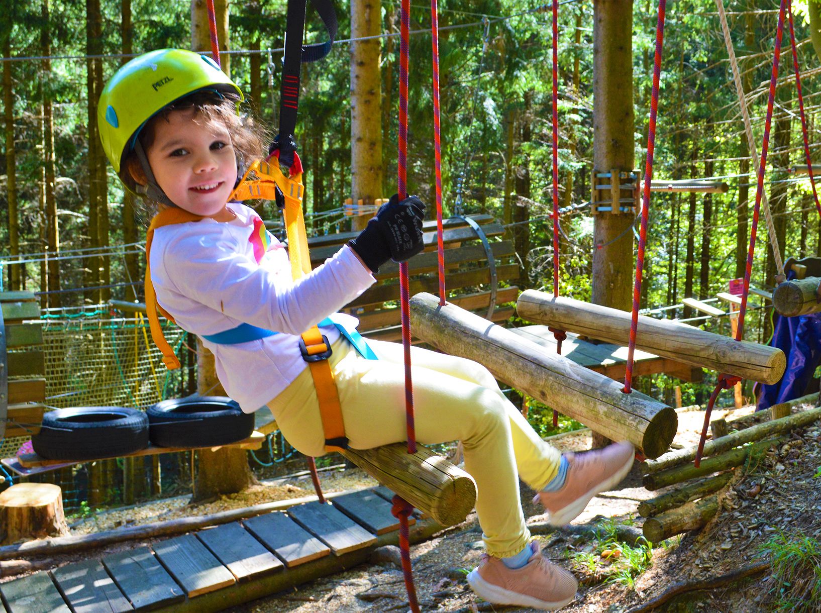 Ein Kind mit Helm und Sicherheitsgurt sitzt auf einem Holzparcours im Wald.