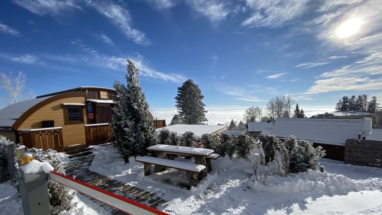 Winterliche Glamping-H&uuml;tte mit Schnee bedeckt, umgeben von B&auml;umen und blauem Himmel.