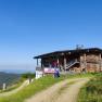 Eine Bergh&uuml;tte aus Holz mit einem roten Fahrzeug davor, umgeben von gr&uuml;nen Wiesen und einem klaren blauen Himmel.