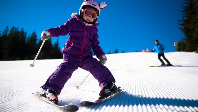 A child in a purple ski suit is skiing on a groomed slope, followed by an adult in a blue suit.