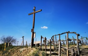 Holzkreuz auf einem H&uuml;gel mit Weinreben und blauem Himmel im Hintergrund.