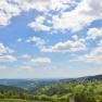 Panoramic view of a hilly landscape with forests and meadows under a blue sky with white clouds.