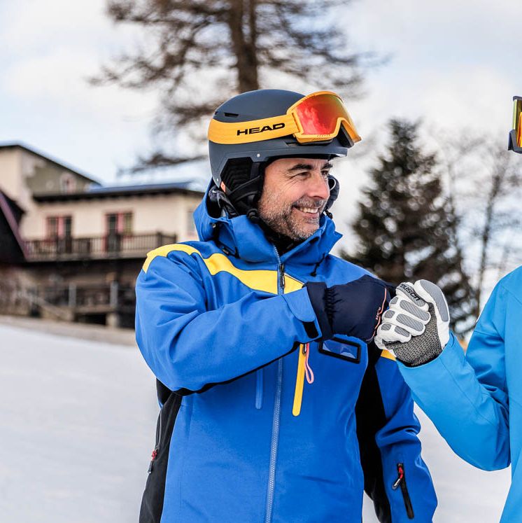 Zwei Personen stehen oder fahren auf einer Skipiste und begrüßen beziehungsweise feiern sich mit einem Fist Bump. Die Szene zeigt einen freundschaftlichen Moment im Skigebiet.