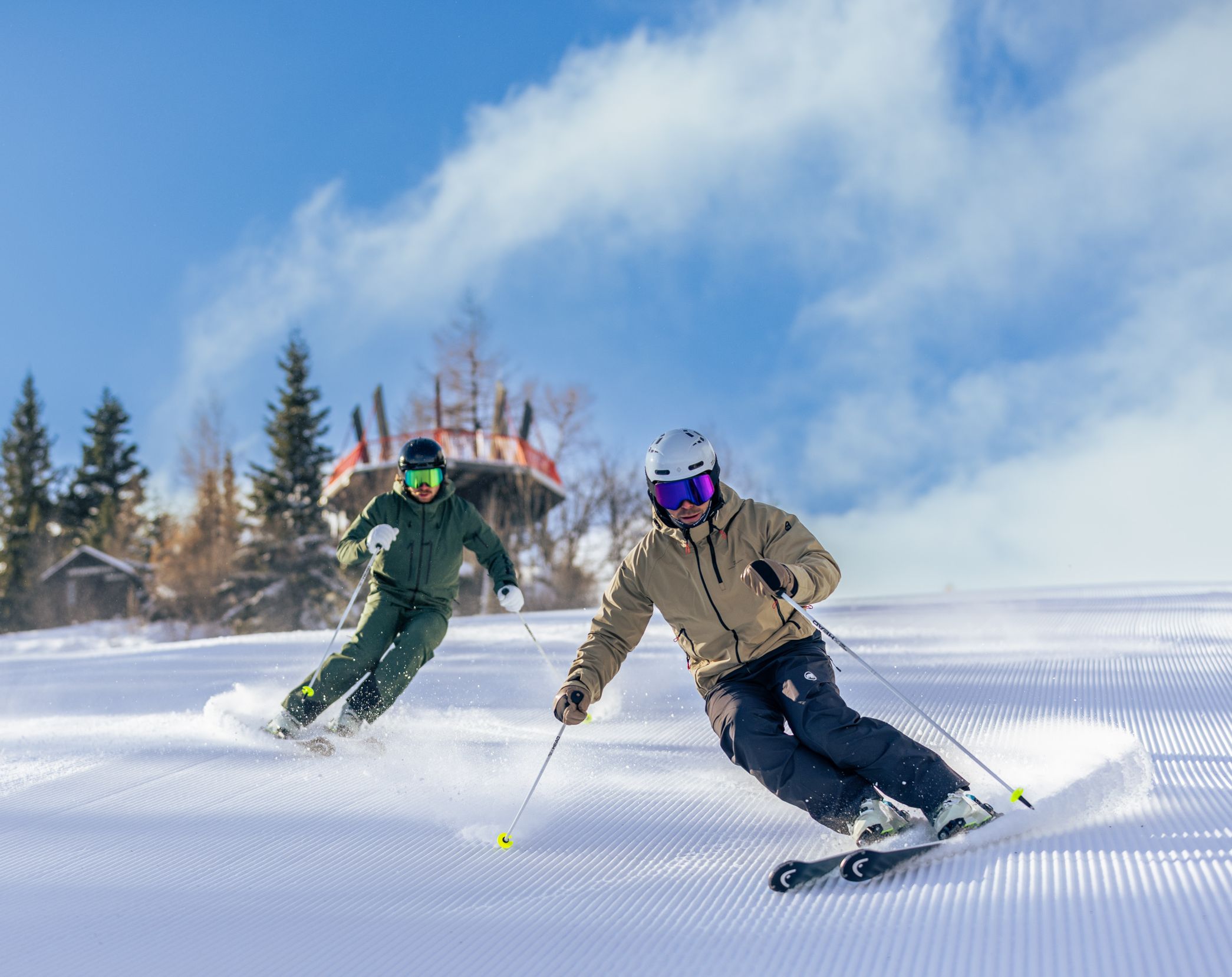 Zwei Männer fahren mit Skiern in schnellen Wedelschwüngen eine präparierte Skipiste hinunter. Die winterliche Umgebung und die verschneite Piste sind im Hintergrund zu sehen.