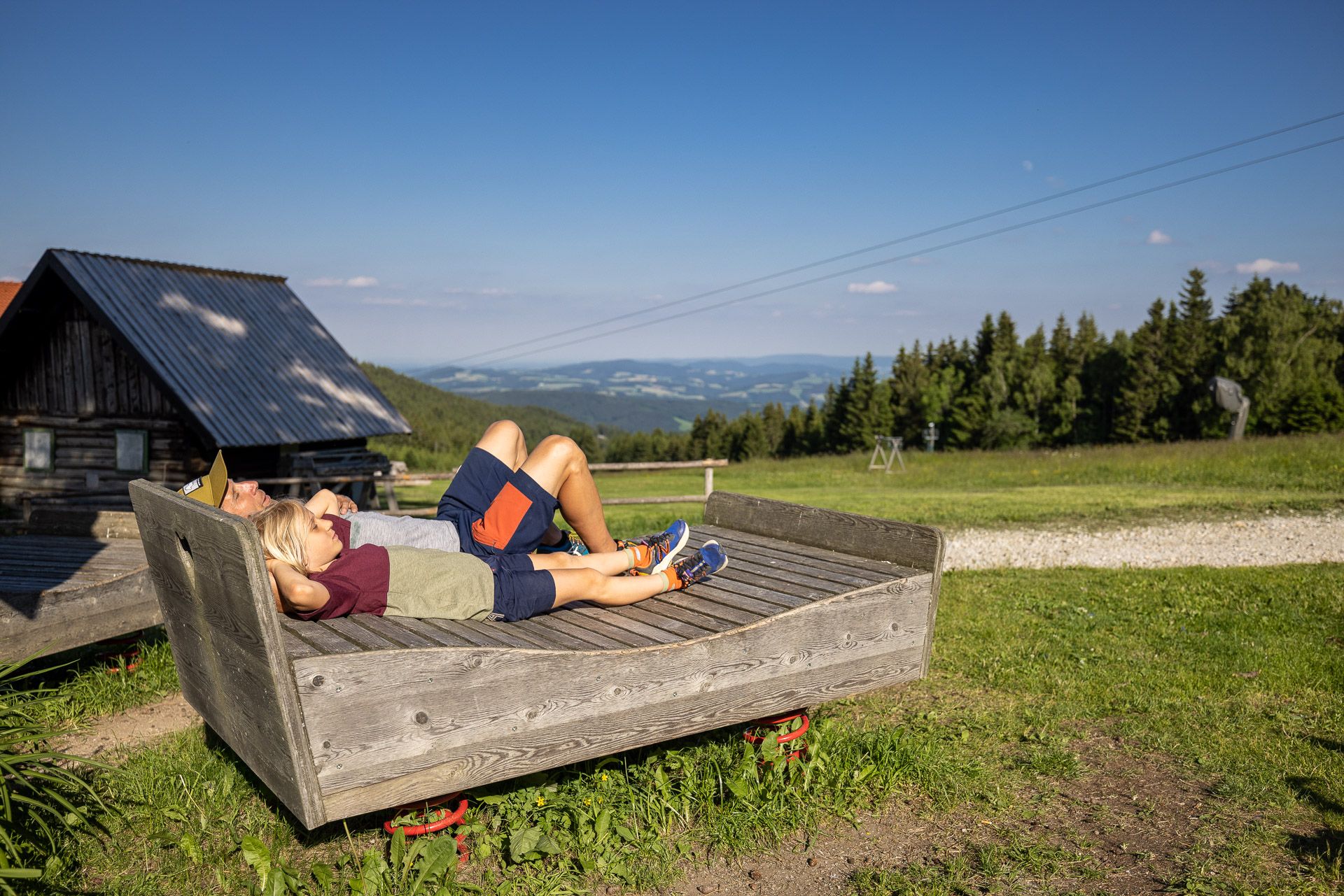 Ein Vater und sein Sohn liegen gemeinsam auf einem Holzbett im Freien und entspannen sich. Im Hintergrund ist bei sonnigem Wetter eine weite Aussicht auf die Landschaft zu sehen.