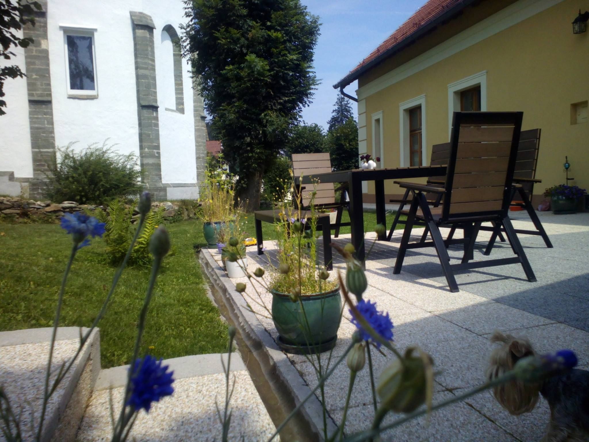 Terrasse mit Tisch, Stühlen und Pflanzen in einem Garten neben einem gelben Haus.