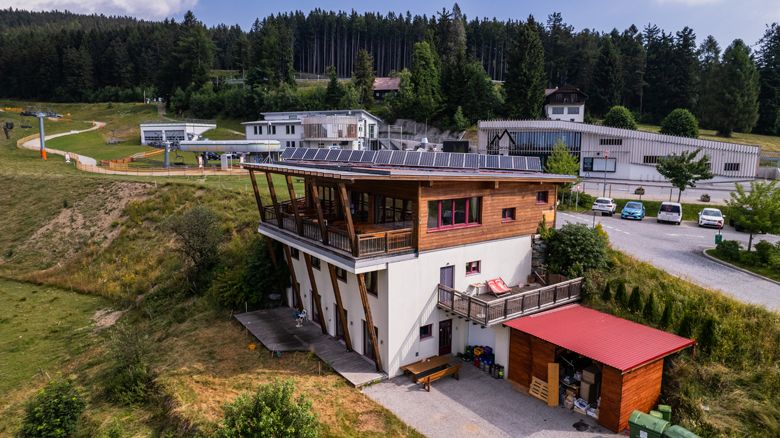 Ein modernes Gebäude mit Solarpanelen auf dem Dach, umgeben von grüner Landschaft und einem Parkplatz mit Autos.