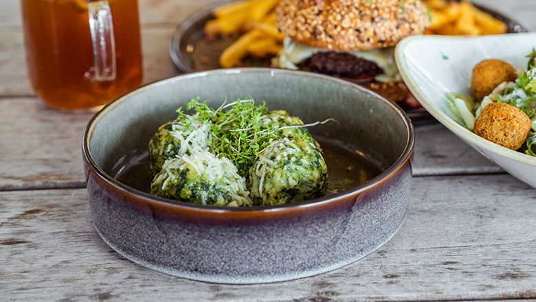 Ein Teller mit Spinatknödeln, Parmesan und Kresse. Im Hintergrund ein Getränk, ein Burger und ein Salat. 