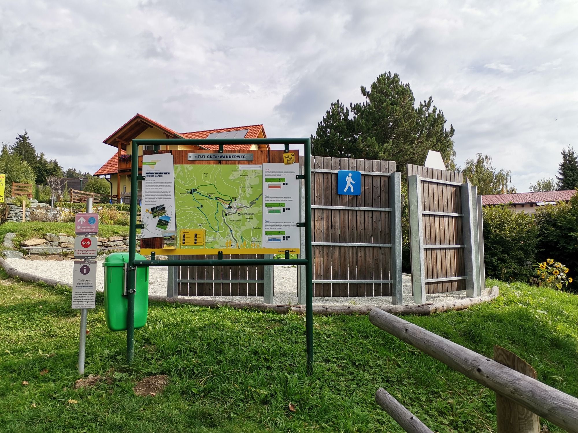 Wanderweg-Infotafel mit Karte und Hinweisschildern auf einer Wiese, umgeben von Bäumen und einem Gebäude im Hintergrund.