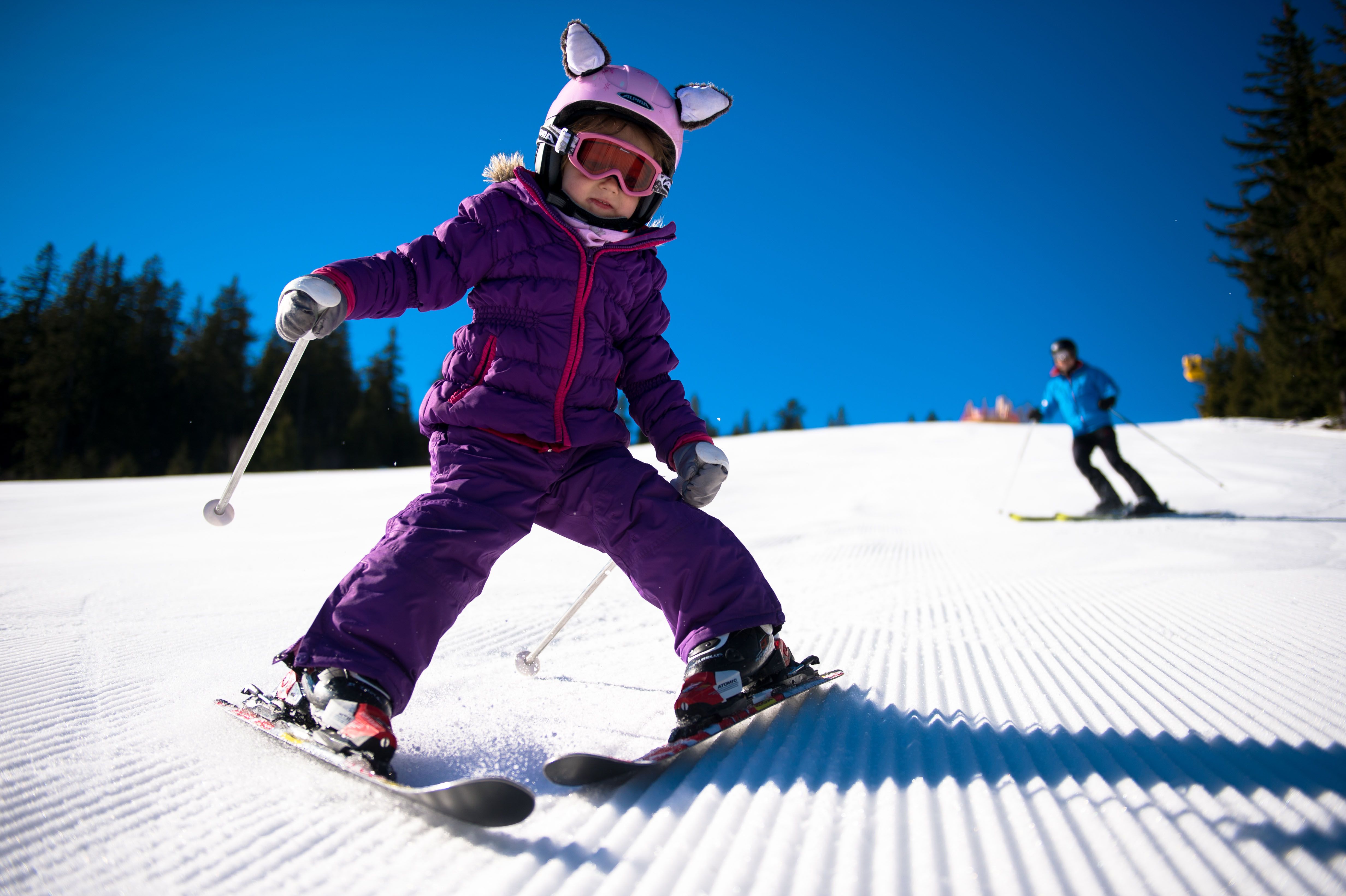 Ein Kind in lila Skianzug fährt auf einer präparierten Piste Ski, gefolgt von einem Erwachsenen im blauen Anzug.