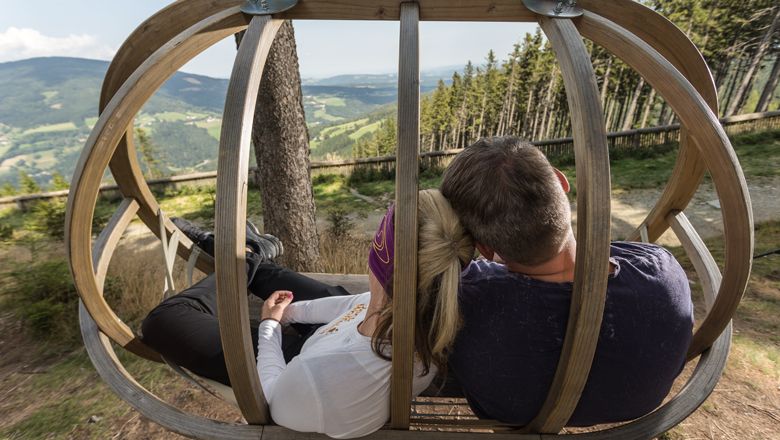 Zwei Personen sitzen in einer Holzschaukel mit Blick auf eine hügelige Landschaft.
