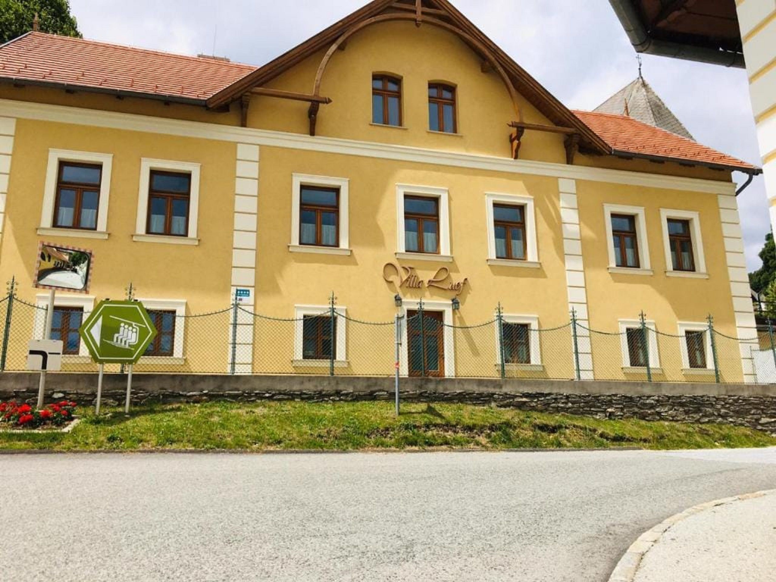 Gelbes Gebäude mit der Aufschrift 'Villa Luef', umgeben von einem Zaun und einer Straße im Vordergrund.