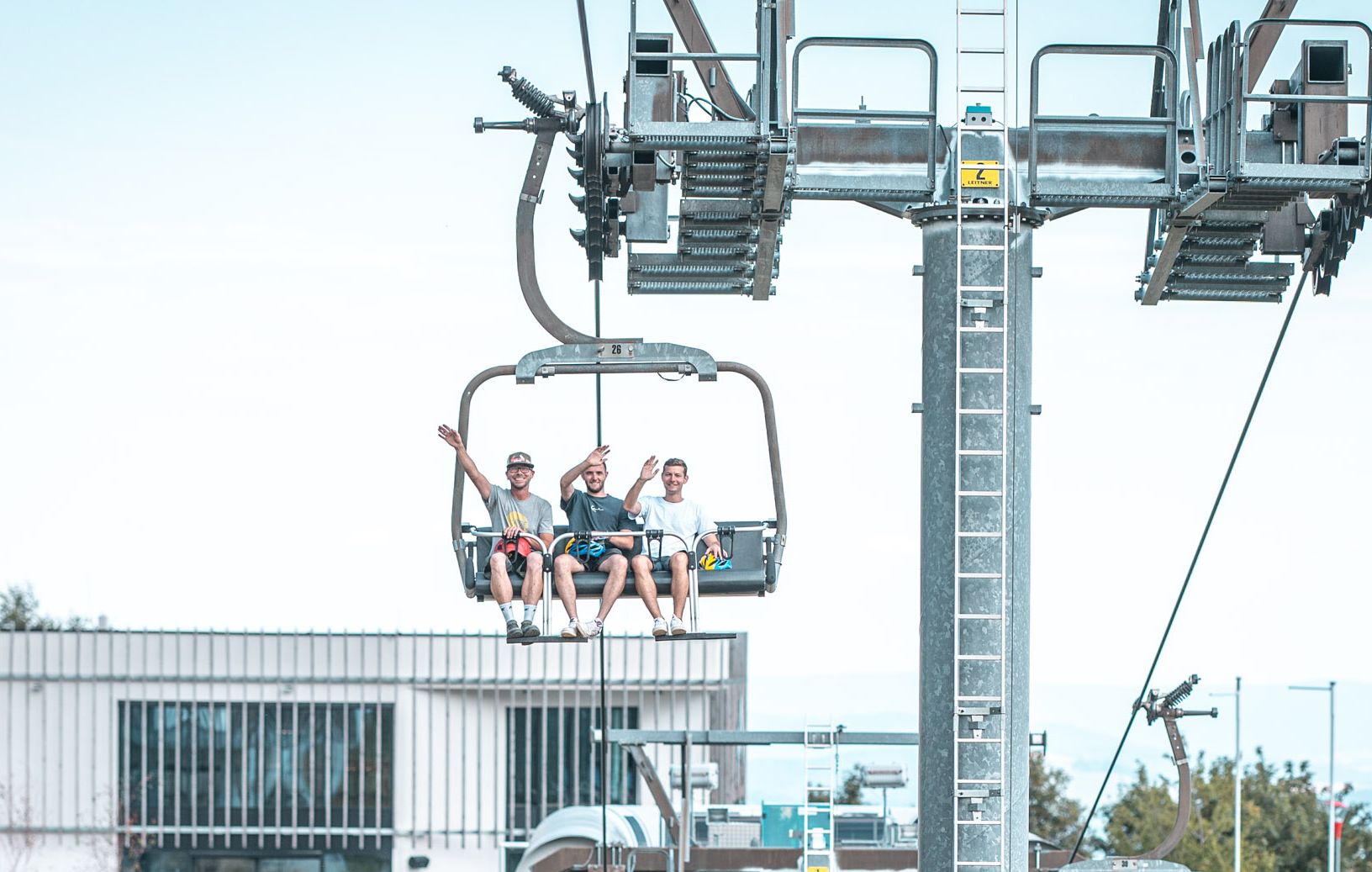 Drei Freunde sitzen gemeinsam auf einem Sessellift und fahren den Berg hinauf. Während der Fahrt winken sie in die Kamera und genießen die gemeinsame Zeit im Freien.