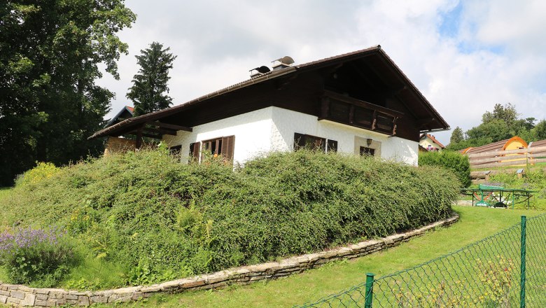 A white house with a wooden roof and garden, surrounded by bushes and trees, under a cloudy sky.