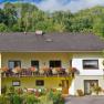Two-storey yellow house with balcony and flowers, surrounded by trees and garden.