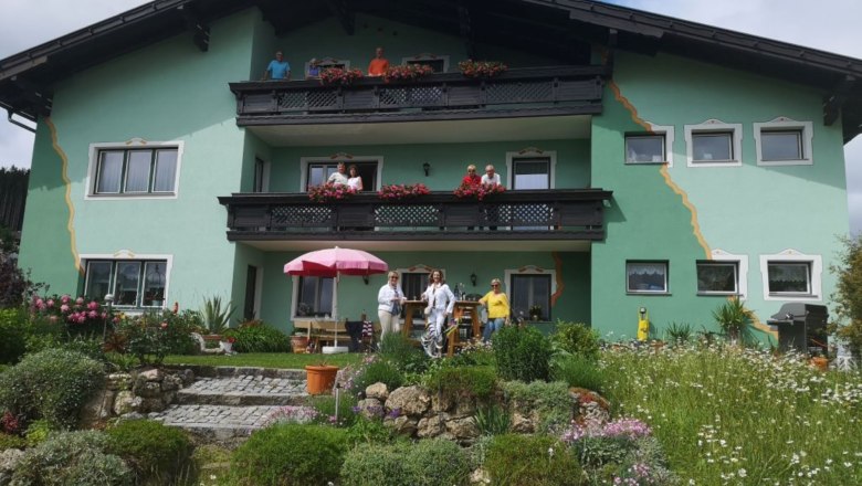 Ein gr&uuml;nes Haus mit Balkonen und Menschen davor, umgeben von einem bl&uuml;henden Garten.