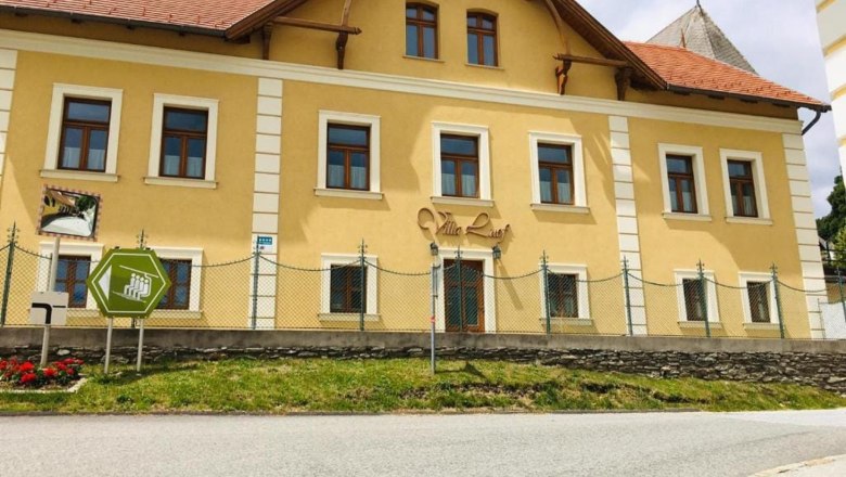 Gelbes Gebäude mit der Aufschrift 'Villa Luef', umgeben von einem Zaun und einer Straße im Vordergrund.