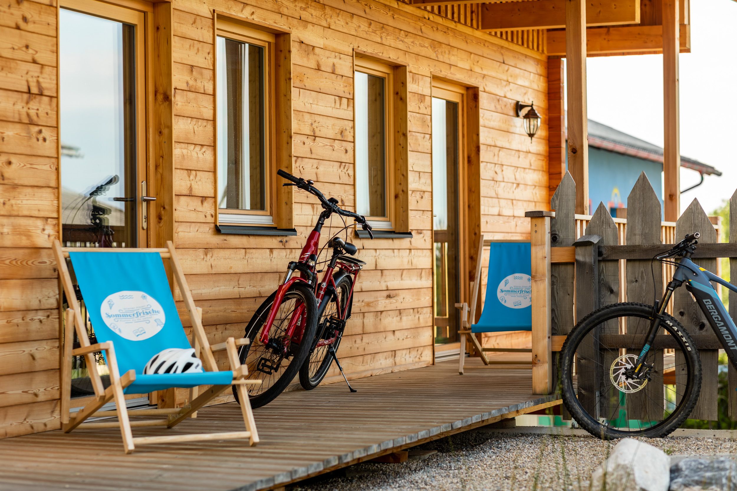 Holzveranda mit zwei Fahrrädern und blauen Liegestühlen vor einem Holzhaus.