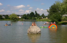 Kinder spielen im Wasser eines Teichs mit bunten Schwimmreifen und einem gro&szlig;en Stein.