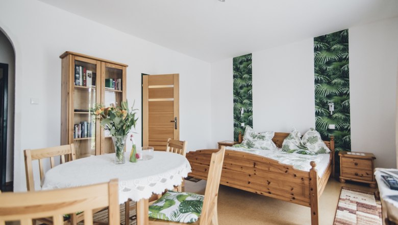 Ein gemütliches Doppelzimmer mit Holzmöbeln, einem Tisch mit Blumen, einem Bett mit Pflanzenmuster und einem Bücherregal.