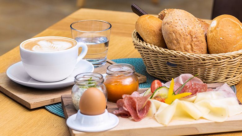 Frühstück auf einem Holzbrett, daneben ein Gebäckkorb und ein Kaffee mit Wasser.