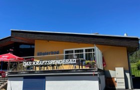 Gelbes Geb&auml;ude mit der Aufschrift 'K&ouml;glerbad Das Kraftspendebad' und Sonnenschirmen auf der Terrasse.