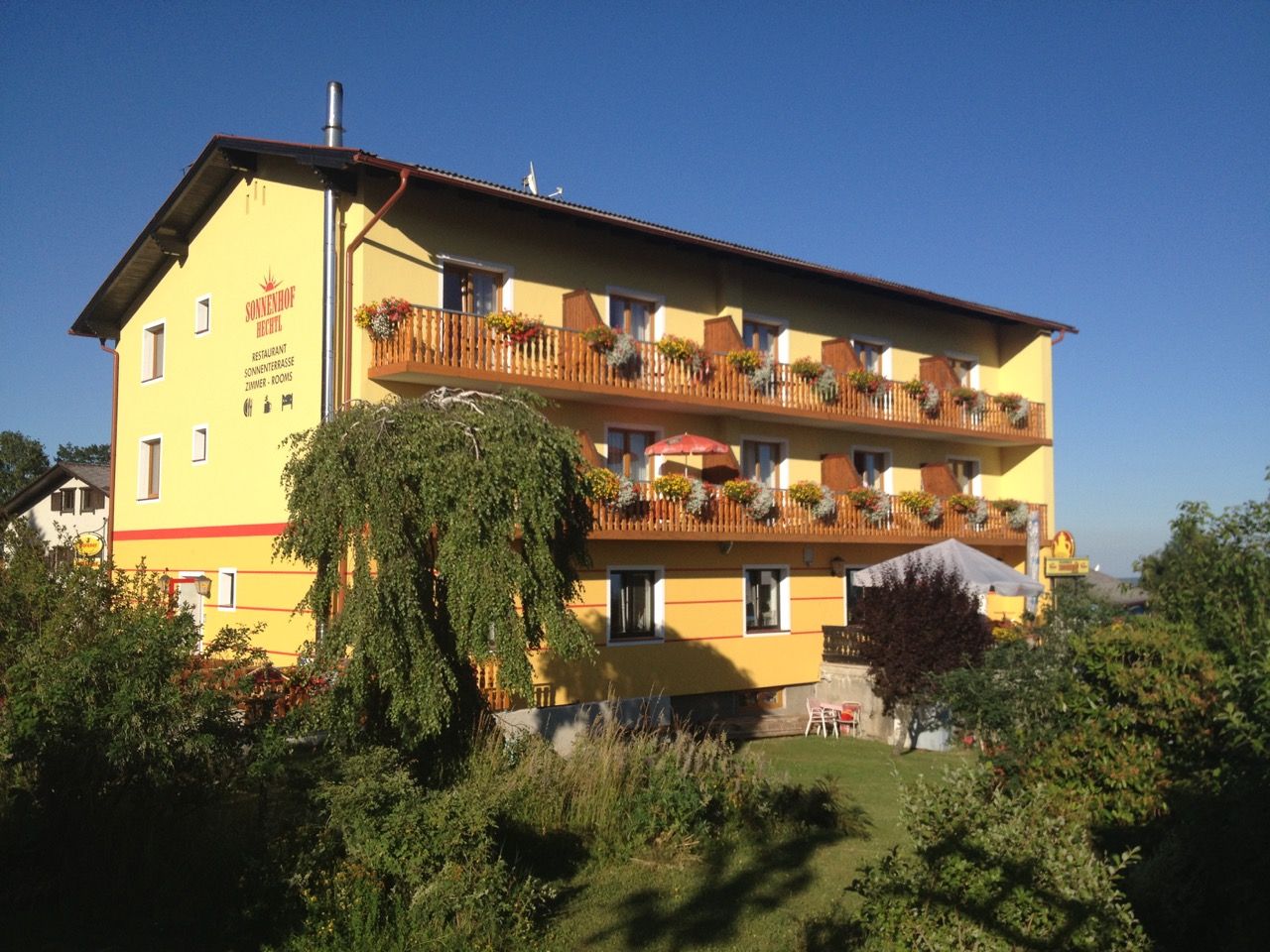 Gelbes Gebäude mit Holzbalkonen und Blumen, umgeben von Bäumen und Garten, unter blauem Himmel.