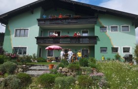 Ein gr&uuml;nes Haus mit Balkonen und Menschen davor, umgeben von einem bl&uuml;henden Garten.