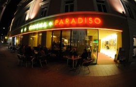 Au&szlig;enansicht eines Kinos namens Cinema Paradiso bei Nacht mit beleuchtetem Eingang und Menschen an Tischen.
