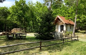 Eine kleine Kapelle mit rotem Dach in einem Waldgebiet, umgeben von B&auml;umen und einem Holzzaun.
