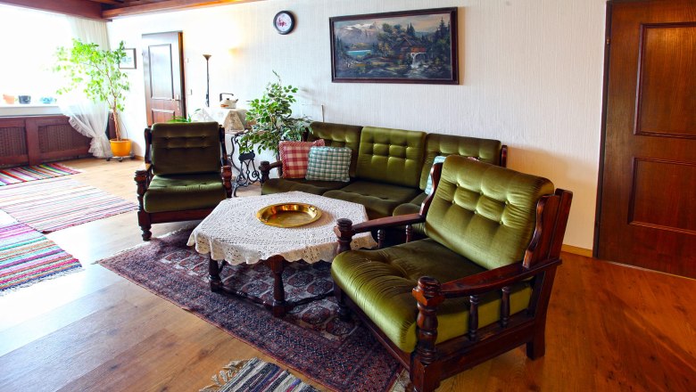 Living room with green upholstered furniture, wooden table with lace ceiling, plants and mural.