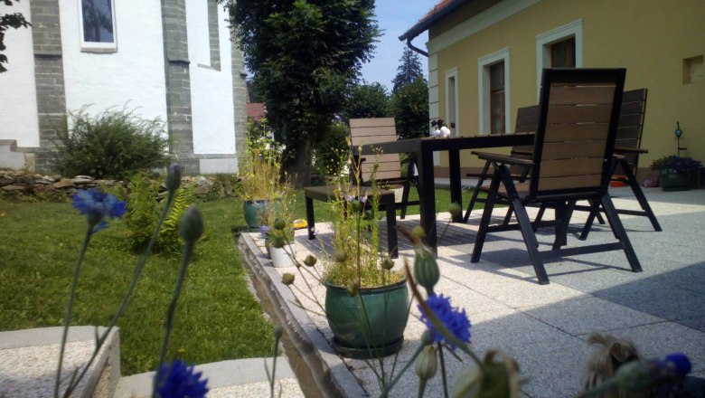 Terrace with table, chairs and plants in a garden next to a yellow house.