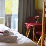Ein gem&uuml;tliches Zimmer mit einem Bett, einem Tisch mit roten St&uuml;hlen und einem Blumenstrau&szlig;. Ein Fenster zeigt einen Balkon und herbstliche B&auml;ume.