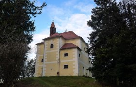 Kapelle mit gelber Fassade auf einem H&uuml;gel, umgeben von B&auml;umen.